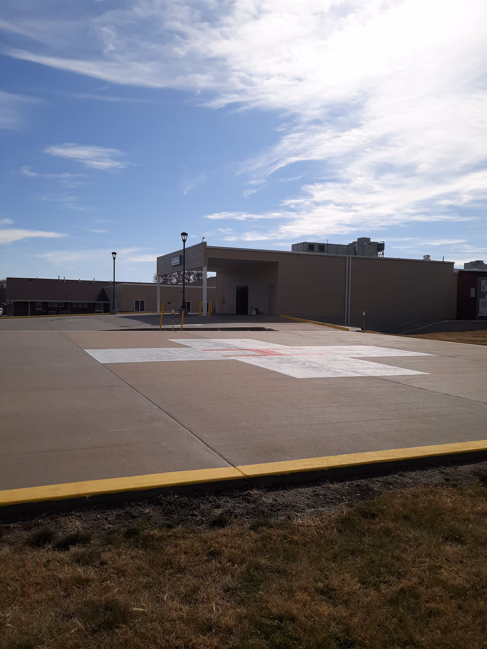 Outdoor view of a building with a large concrete area featuring a white and red cross painted on the ground, likely a helipad, under a partly cloudy sky.