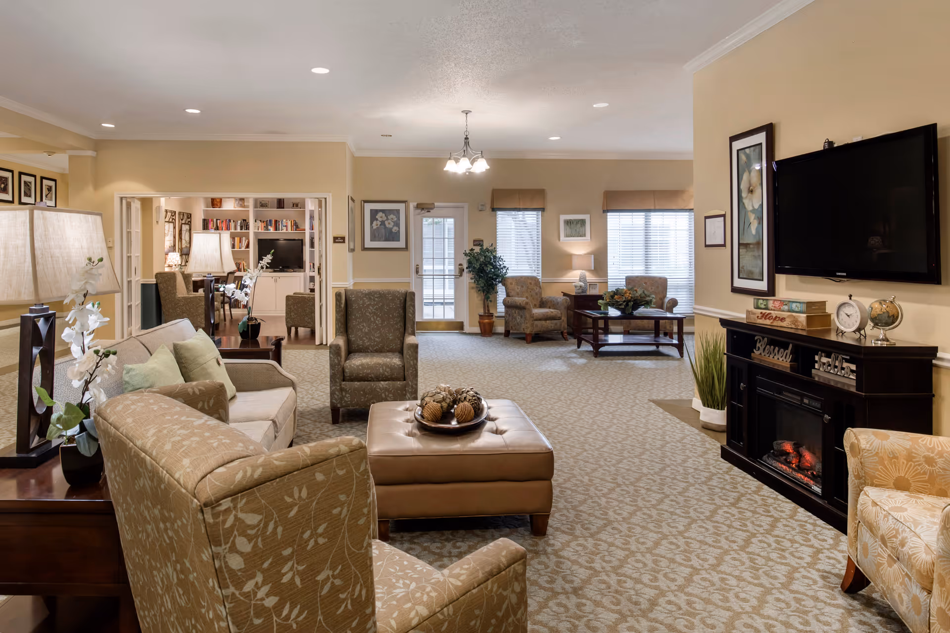 A cozy senior living facility common area with beige walls and patterned carpet. The room features several upholstered armchairs and a sofa arranged around a leather ottoman with decorative pinecones. There is a dark wood electric fireplace with a flat-screen TV mounted above it. The room is well-lit with ceiling lights and table lamps, and there are framed floral artworks on the walls. In the background, a doorway leads to a library area with bookshelves and additional seating.