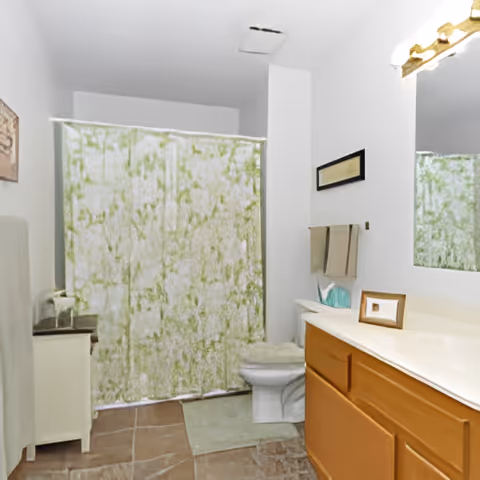 A bathroom with a green and white patterned shower curtain, a white toilet, a wooden vanity with a white countertop, a large mirror above the vanity, a small framed picture on the countertop, and a towel rack with two beige towels. The floor is tiled in a brownish color.