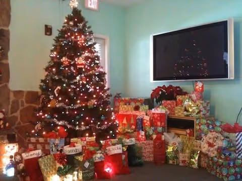 A decorated Christmas tree with colorful lights and ornaments stands in a room next to a stone wall. Beneath the tree and nearby are numerous wrapped Christmas presents in various sizes and festive wrapping paper. A large flat-screen TV is mounted on the wall above a small fireplace, and a poinsettia plant is placed on the fireplace mantel.