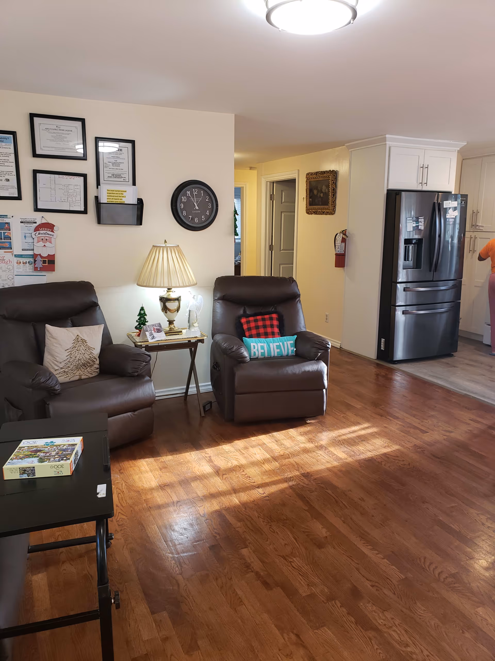 Cozy living room with two brown recliners, a side table and lamp, wall clock and framed certificates, and a view into a kitchen with a stainless steel refrigerator.