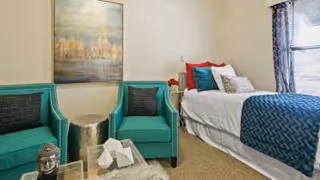 A cozy bedroom featuring a single bed with white and teal bedding, two teal armchairs with a small metallic side table between them, a large painting on the wall, and a window with patterned curtains allowing natural light into the room.