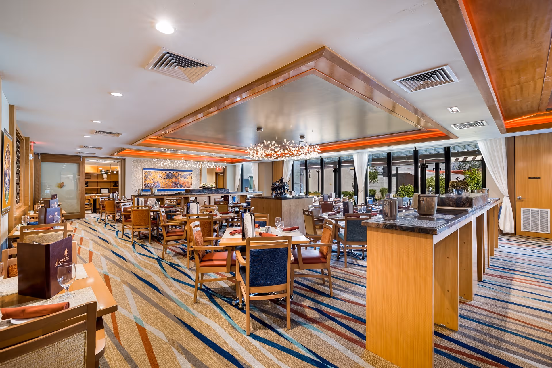 Spacious dining room with multiple wooden tables and chairs arranged neatly. The room features large windows with white curtains allowing natural light to brighten the space. Modern chandeliers hang from the ceiling, and the carpet has a colorful striped pattern. Menus and glassware are set on the tables, and there is a long counter with decorative items on the right side.