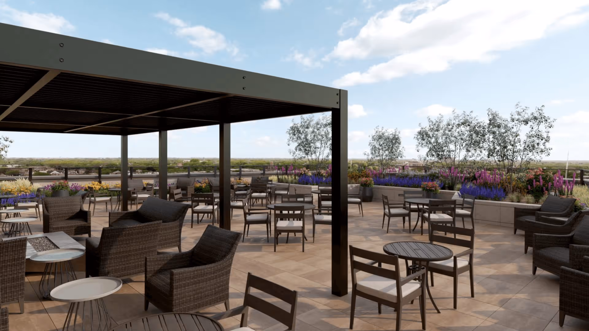 Outdoor patio area with multiple tables and chairs, some under a pergola, surrounded by planters with colorful flowers and small trees, under a partly cloudy sky.