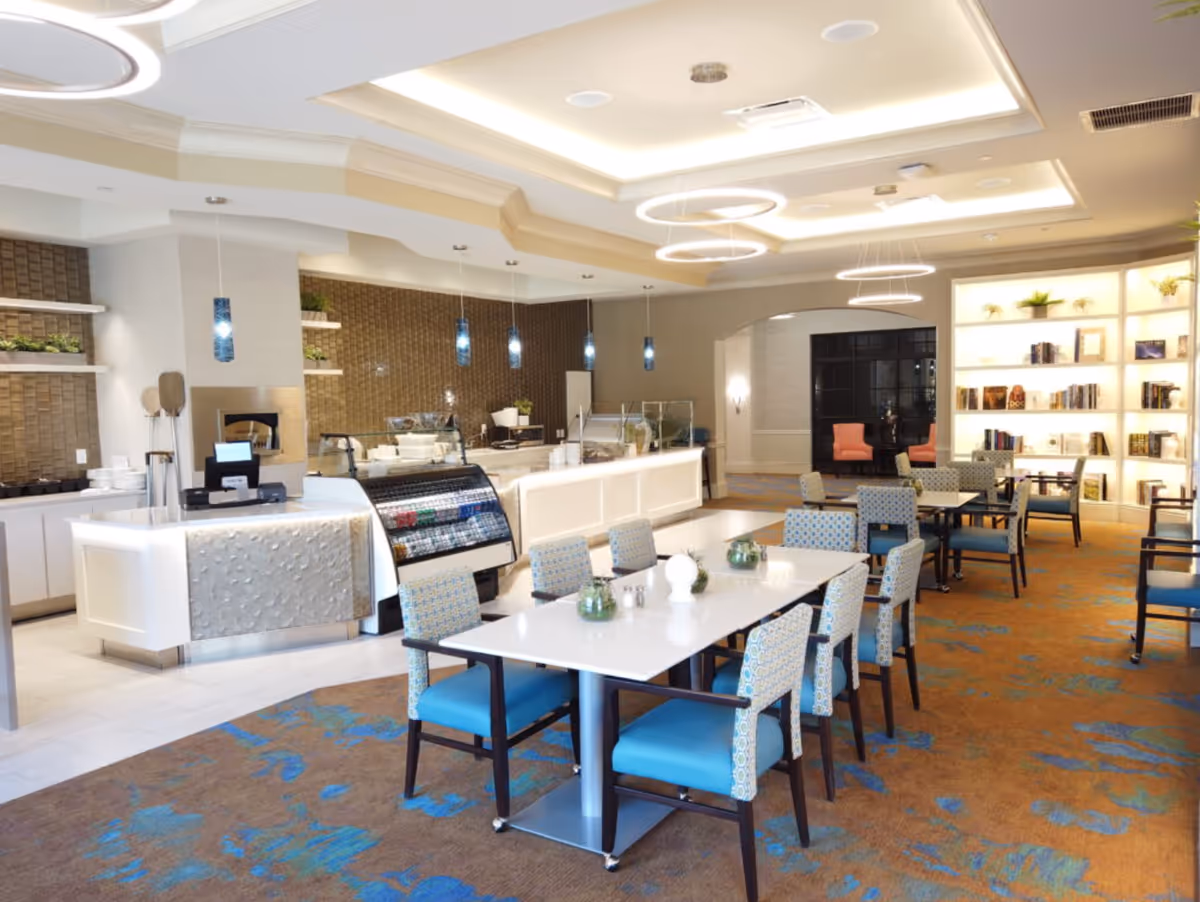 A bright and modern dining area in a senior living facility featuring several tables with blue cushioned chairs. The room has a buffet counter with a glass display and hanging pendant lights. There is a bookshelf with decorative plants and books in the background, and the ceiling has recessed lighting with circular light fixtures.