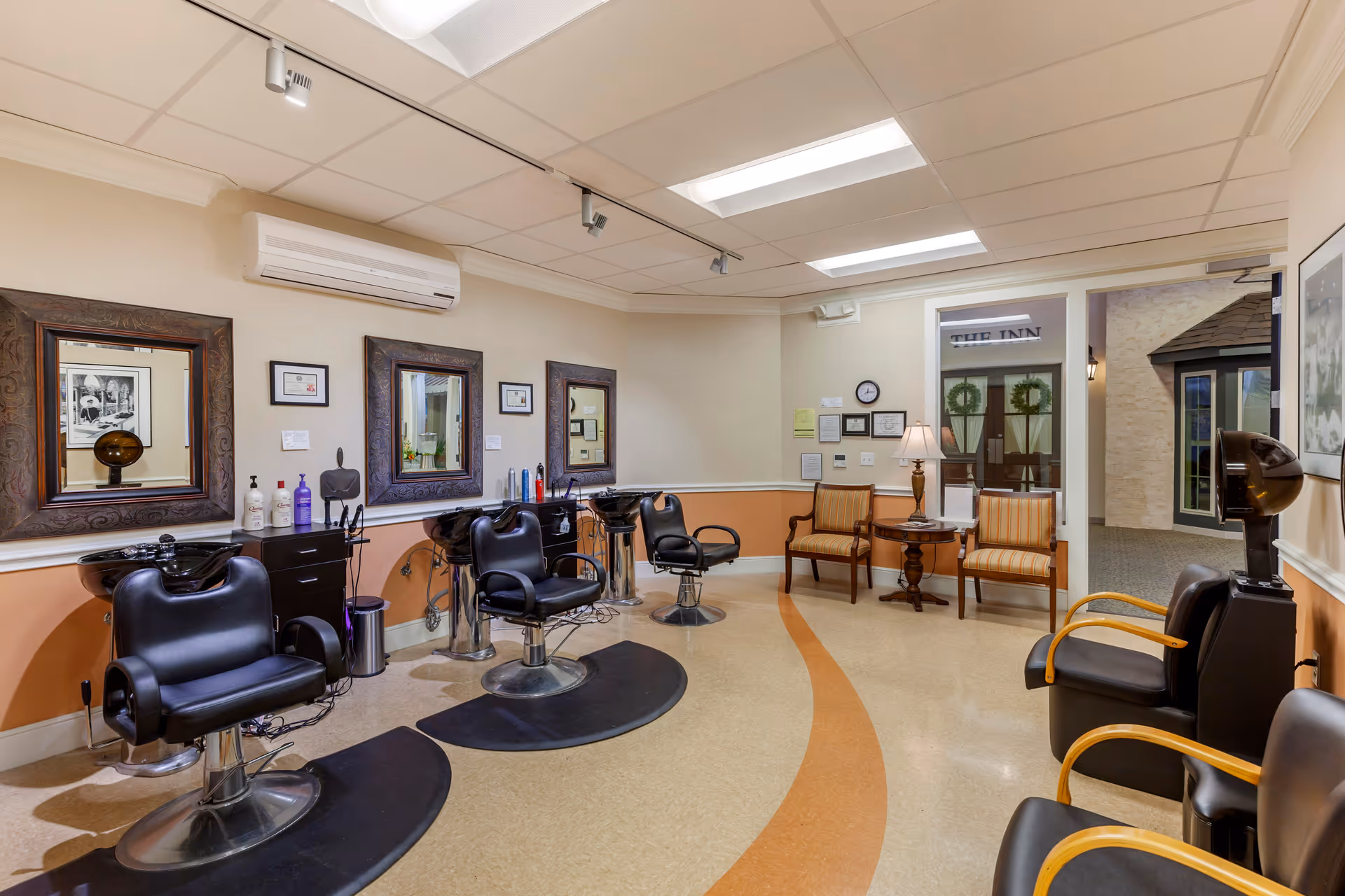 Interior view of a hair salon area in a senior living facility with three black salon chairs in front of mirrors on the left side, hair washing sinks, and a seating area with two striped armchairs and a small table with a lamp on the right side. The room has a beige and peach color scheme with overhead fluorescent lighting.
