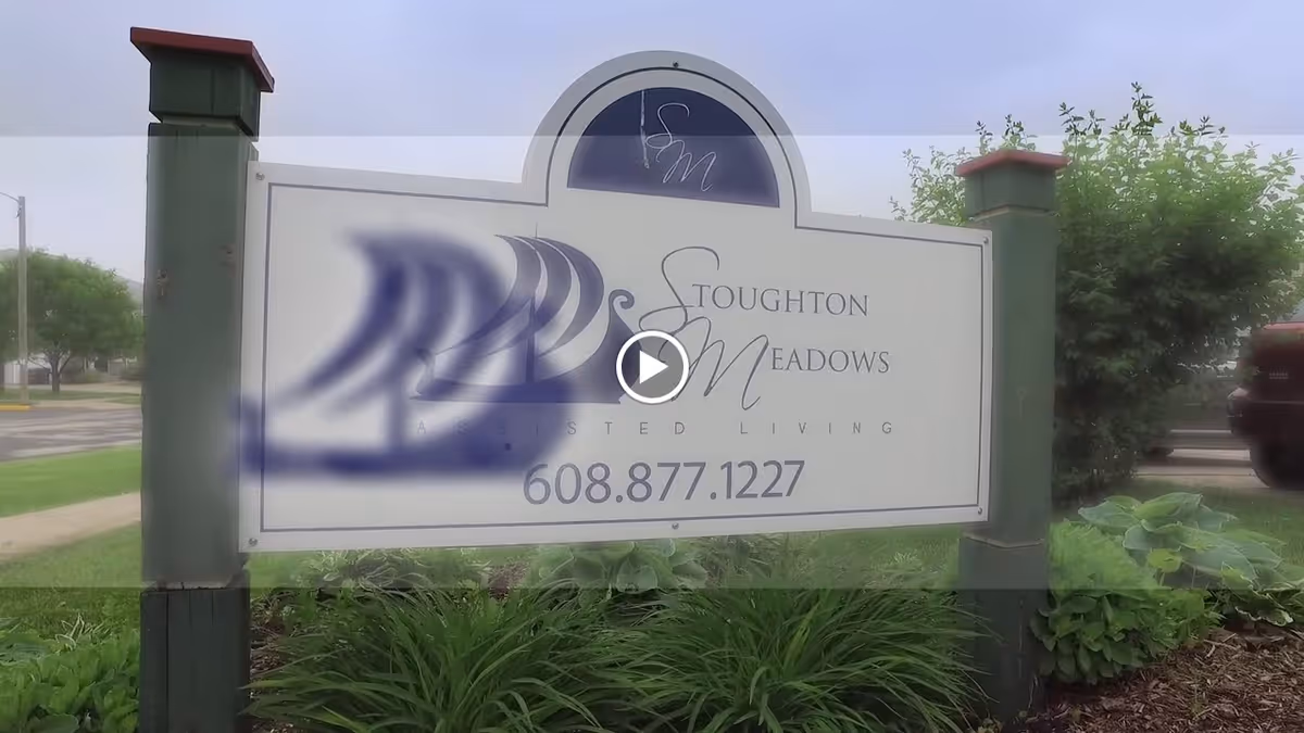 Outdoor sign reading "Stoughton Meadows Assisted Living" with a phone number mounted between posts in a landscaped front yard.