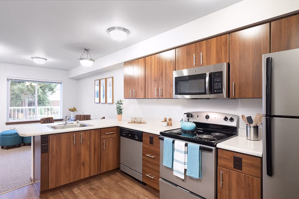 Modern kitchen with wooden cabinets, stainless steel appliances including a refrigerator, microwave, stove with a teal kettle, and dishwasher. The kitchen has a white countertop with a sink and two blue towels hanging on the oven handle. In the background, there is a window with a view of trees and part of a living area with chairs and framed artwork on the wall.