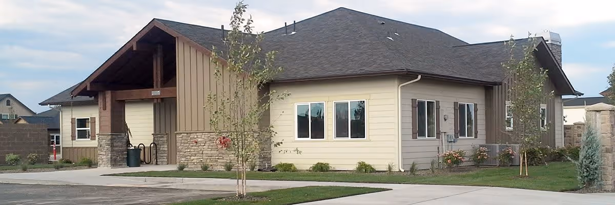 Exterior view of a single-story building with beige siding, stone accents, and a dark shingled roof. The building has several windows and a covered entrance supported by wooden beams. There are small trees and shrubs planted around the building, and a paved driveway and sidewalk in the foreground.