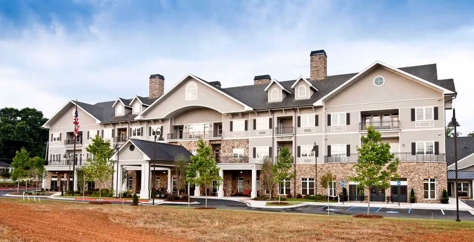 Exterior view of a large, multi-story senior living facility building with beige siding, stone accents, multiple chimneys, and a covered entrance. The building is surrounded by young trees, a parking lot with handicap spaces, and a well-maintained lawn under a partly cloudy sky.