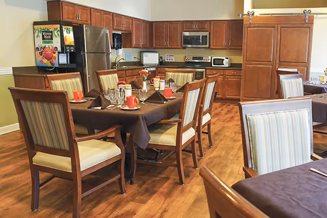 Dining area with wooden tables covered in brown tablecloths, set with glasses, cups, napkins, and condiments. The room has wooden chairs with striped cushions and a kitchen area in the background featuring wooden cabinets, a stainless steel refrigerator, microwave, and a juice dispenser.