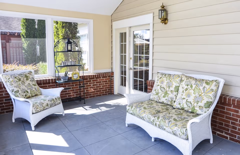 A bright and cozy enclosed patio area with white wicker furniture including a cushioned armchair and loveseat with floral patterned cushions. There is a black metal shelf with decorative items against a half brick wall and large windows letting in natural light. A glass door with white framing leads outside.