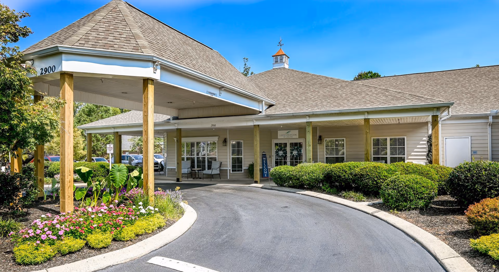 Front entrance and porte-cochere of a senior living building with a curved driveway, seating, and landscaped flowerbeds.