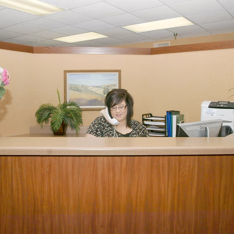 A woman with dark hair and glasses is sitting behind a wooden reception desk, talking on a white corded telephone. The desk has a beige countertop and there is a potted plant with green leaves on it. Behind her, there is a framed landscape painting on a beige wall, some office supplies including a printer, folders, and a computer monitor. The ceiling has fluorescent lights.