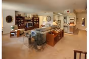 A spacious living room area in a senior living facility featuring beige carpeting, a large gray sofa, wooden bookshelves with a television, framed pictures on the walls, and a small dining area with a table and chairs. The room is well-lit with natural and artificial light, and there is a kitchen area visible in the background.