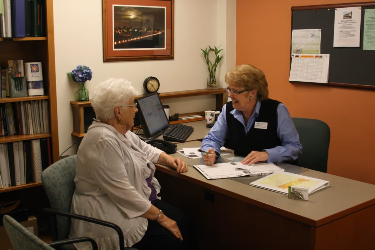 An elderly woman and a staff member are seated at a desk in an office setting. The staff member is smiling and holding a pen while discussing documents with the elderly woman. The office has a computer, bookshelves, a clock, a bulletin board with papers pinned, and some decorative plants and a framed picture on the wall.
