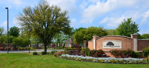 Entrance of The Oaks of Louisiana with a brick sign, landscaped flowerbeds, trees, and a driveway.