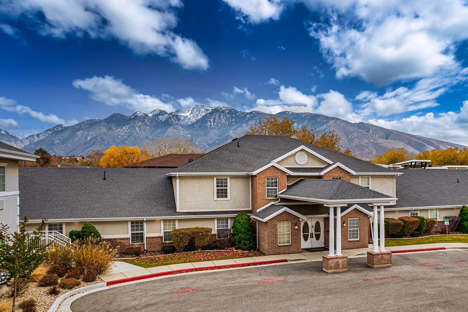 Two-story senior living building with a covered portico entrance and mountains in the background under a blue, partly cloudy sky.