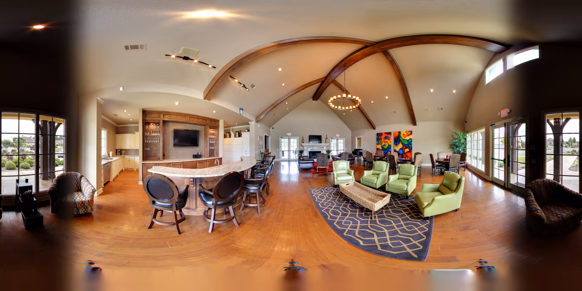 A spacious and well-lit common area in a senior living facility featuring a high vaulted ceiling with wooden beams, a large circular chandelier, and multiple seating arrangements including green armchairs around a patterned rug, bar stools at a curved counter, and additional chairs near large windows and glass doors that provide views of the outdoor area. The room also includes a fireplace, colorful abstract artwork on the wall, and a kitchen area with white cabinetry and a countertop.