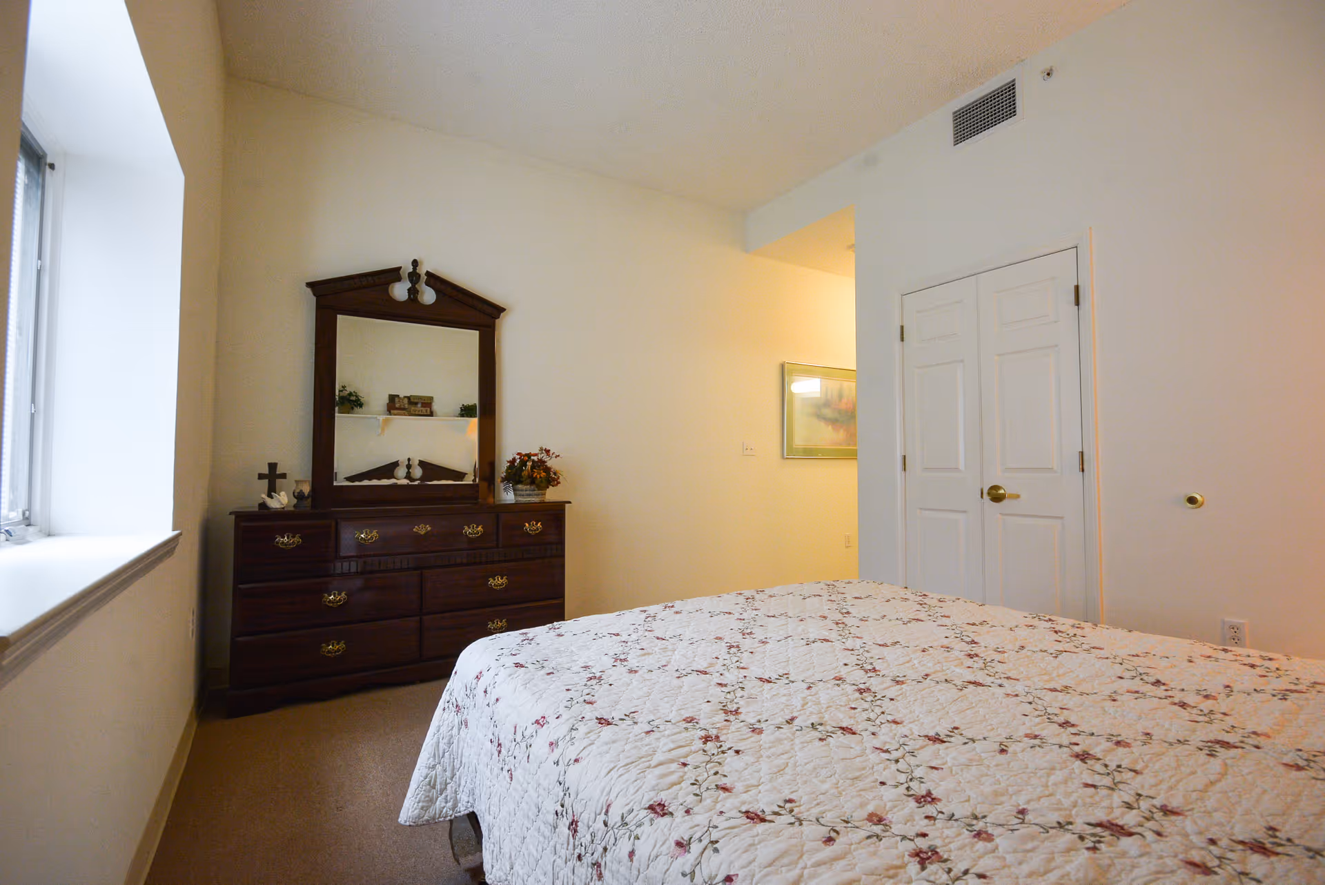 A bedroom in an assisted living facility with a bed covered in a floral quilt, a wooden dresser with a mirror, and a window letting in natural light. There is a closet with double doors and a framed picture on the wall.