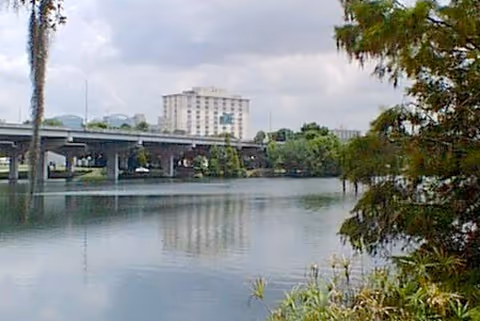 A calm river with a bridge to the left and a multi-story building visible beyond, framed by trees in the foreground.
