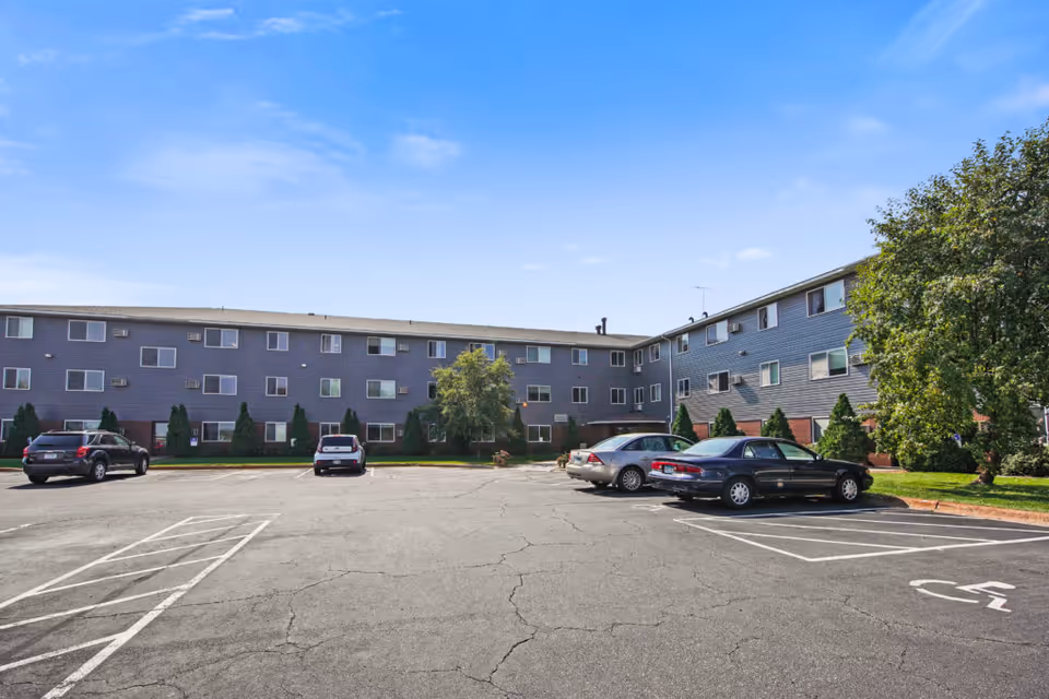 Exterior view of a three-story senior living facility named Keller Lake Commons with a parking lot in front. Several cars are parked, and there are trees and shrubs around the building under a clear blue sky.