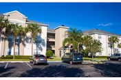 Three-story beige apartment building with palm trees and parked cars in the front parking lot under a clear blue sky.