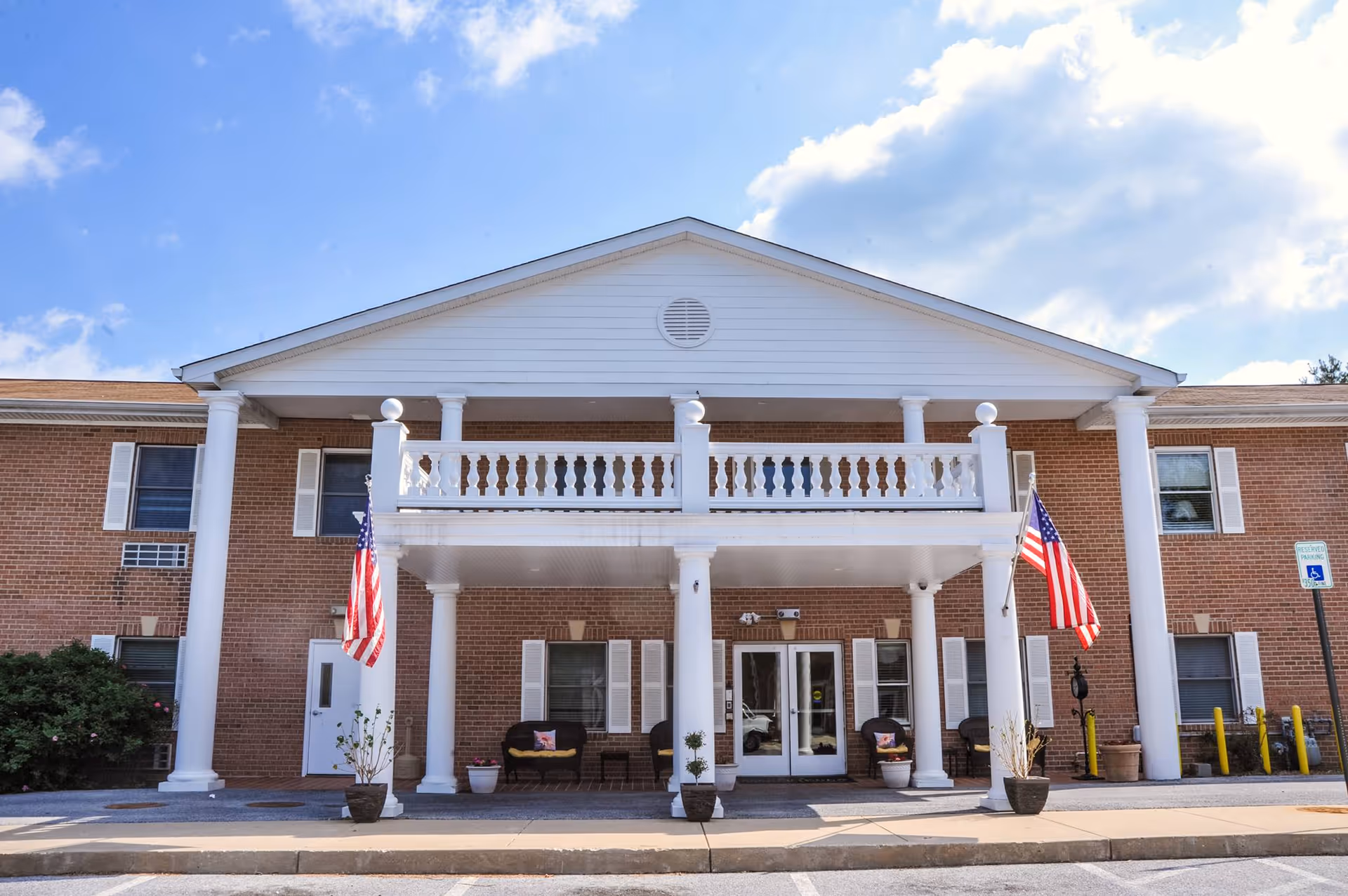 Front entrance of a two-story brick assisted living building with white columns, a balcony, and American flags.