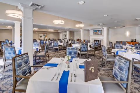 A spacious dining room with multiple tables covered in white tablecloths and blue table runners. Each table is set with silverware, glasses, and small floral centerpieces. The room features patterned carpet, cushioned chairs with blue and gray upholstery, and soft overhead lighting. There is a fireplace and artwork on the walls in the background.
