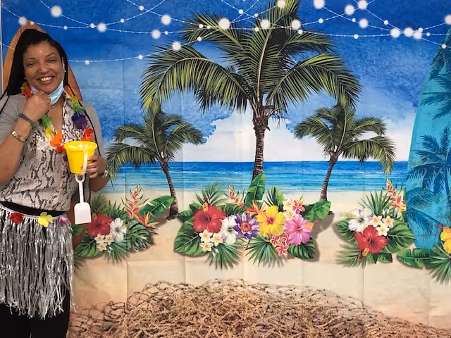 A smiling woman dressed in a tropical-themed outfit stands in front of a beach-themed backdrop with palm trees, flowers, and ocean waves. She is holding a yellow drink and wearing a silver grass skirt and colorful lei.