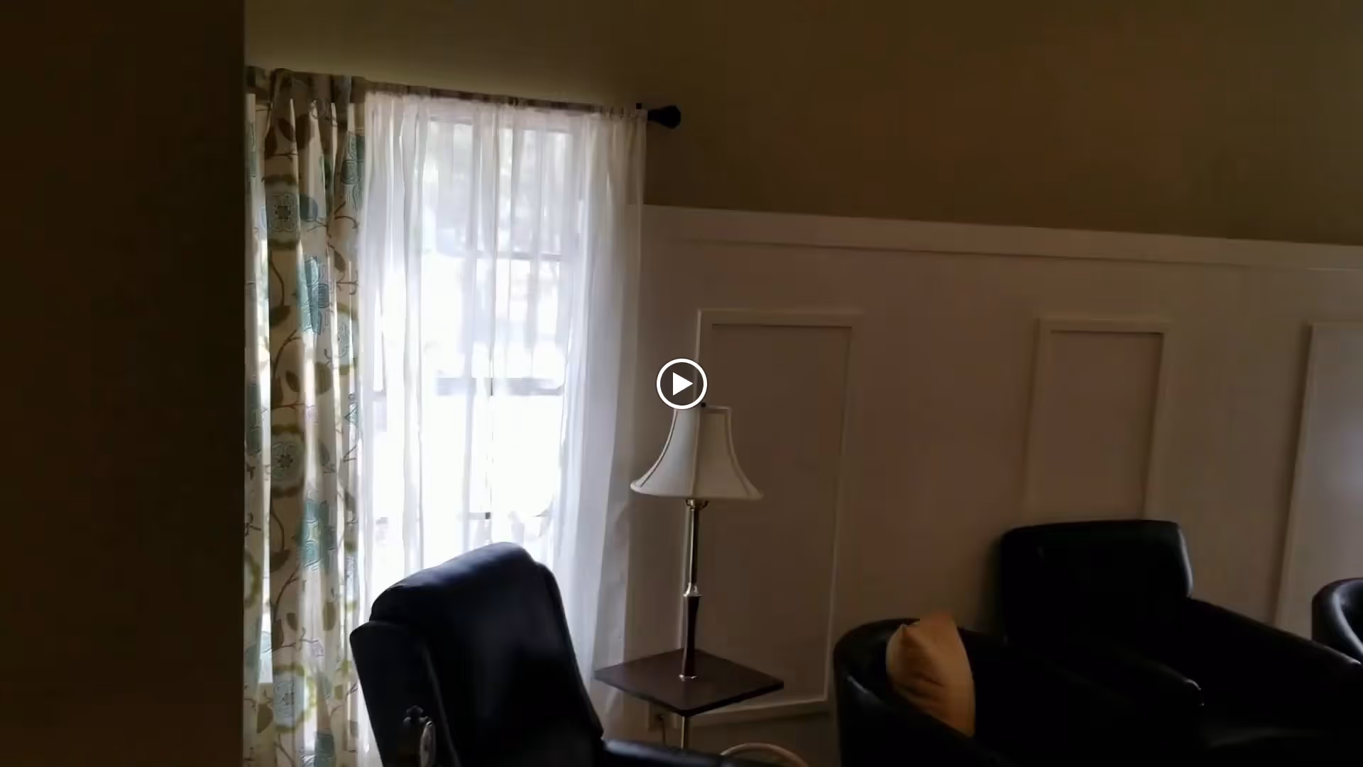 A cozy living room area with two black armchairs, one with a beige pillow, a small wooden side table with a lamp, and a window with sheer white curtains and patterned drapes allowing natural light to enter.