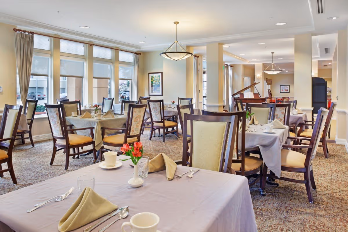 Dining room with neatly set tables and chairs, large windows, and a piano in the background.