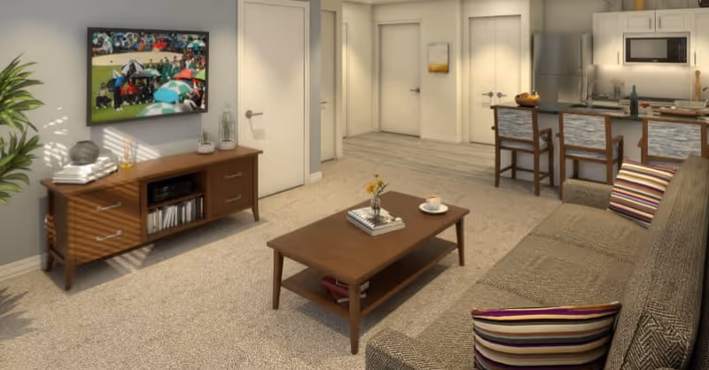 A cozy living room with a brown sofa adorned with striped pillows, a wooden coffee table with a small vase of flowers and a cup, and a wooden TV stand holding books and decorative items. A flat-screen TV is mounted on the wall showing a sports event. The room opens into a kitchen area with a breakfast bar and three stools, featuring stainless steel appliances including a refrigerator and microwave. The walls are light-colored, and the floor transitions from carpet in the living area to wood in the kitchen.