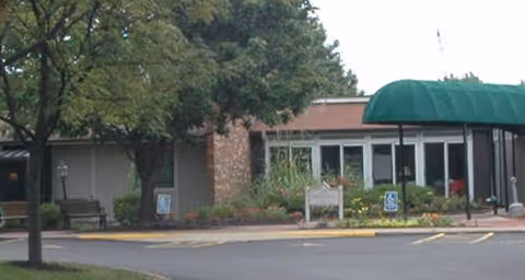 Exterior view of a single-story building with large windows and a green awning over the entrance. There are trees and landscaped plants around the entrance, along with a bench and handicap parking signs visible near the sidewalk.
