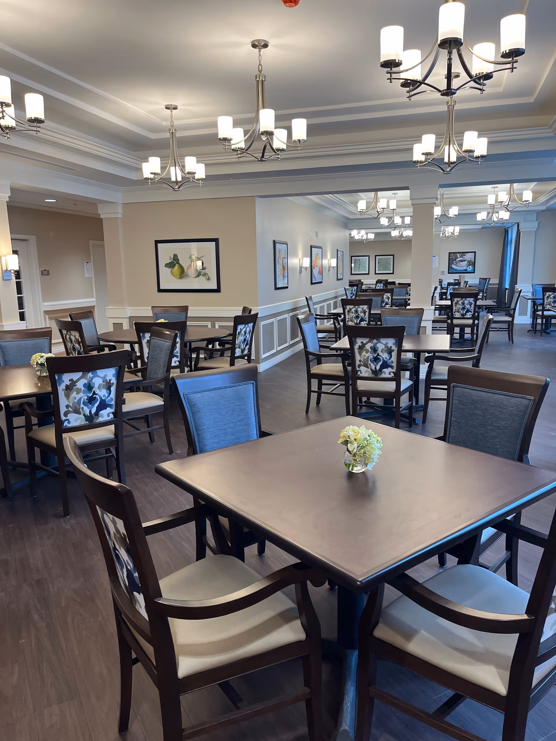 Bright assisted-living dining room with multiple wooden tables and chairs, chandeliers, and small floral centerpieces.