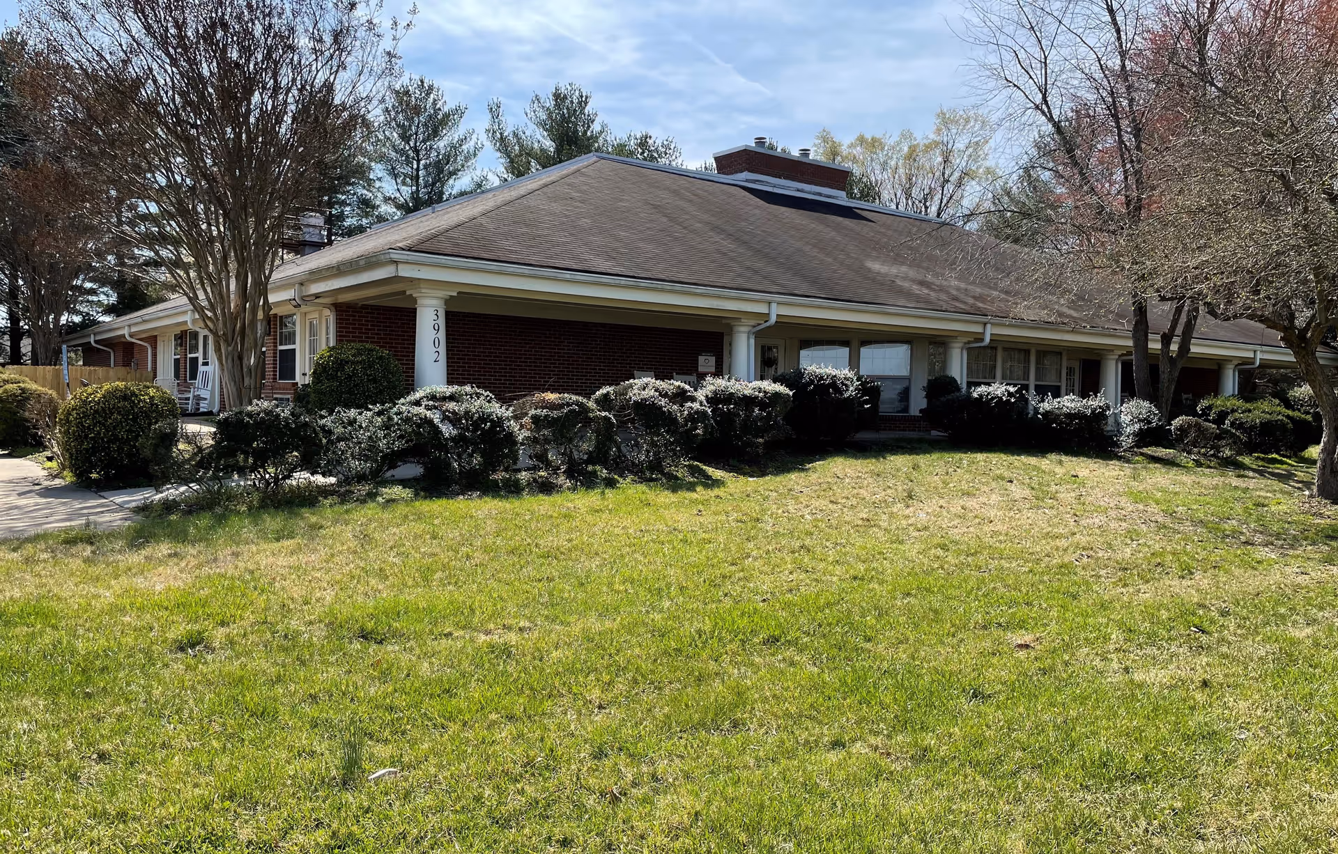 Exterior view of a single-story brick building with a sloped roof, surrounded by bushes and trees, and a grassy lawn in the foreground under a partly cloudy sky.
