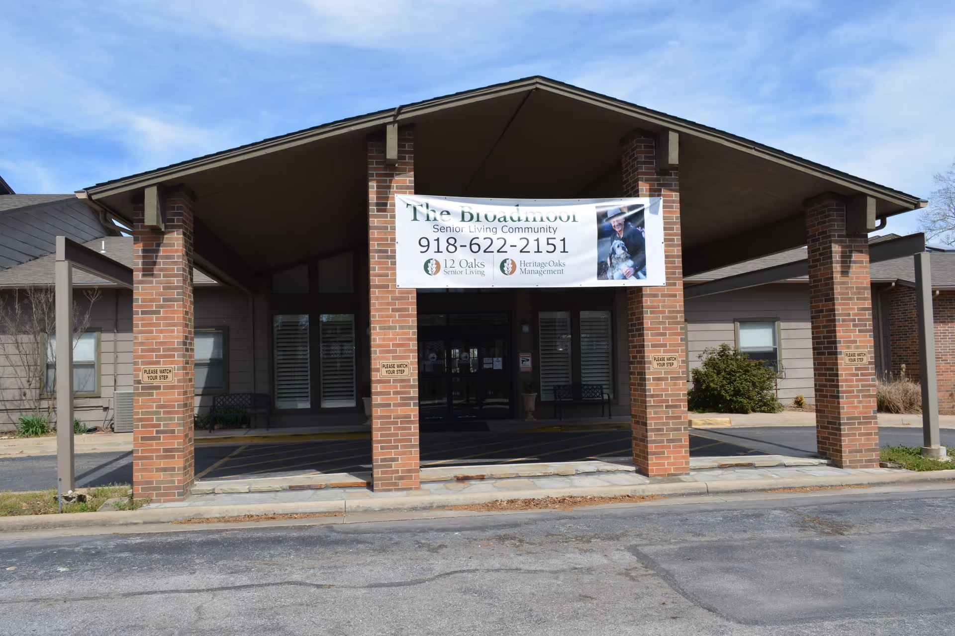 Front entrance of The Broadmoor Senior Living Community featuring a covered drop-off area supported by brick pillars. A banner with the community name, phone number, and management logos hangs above the entrance. The building has a brown exterior with windows and bushes on either side.