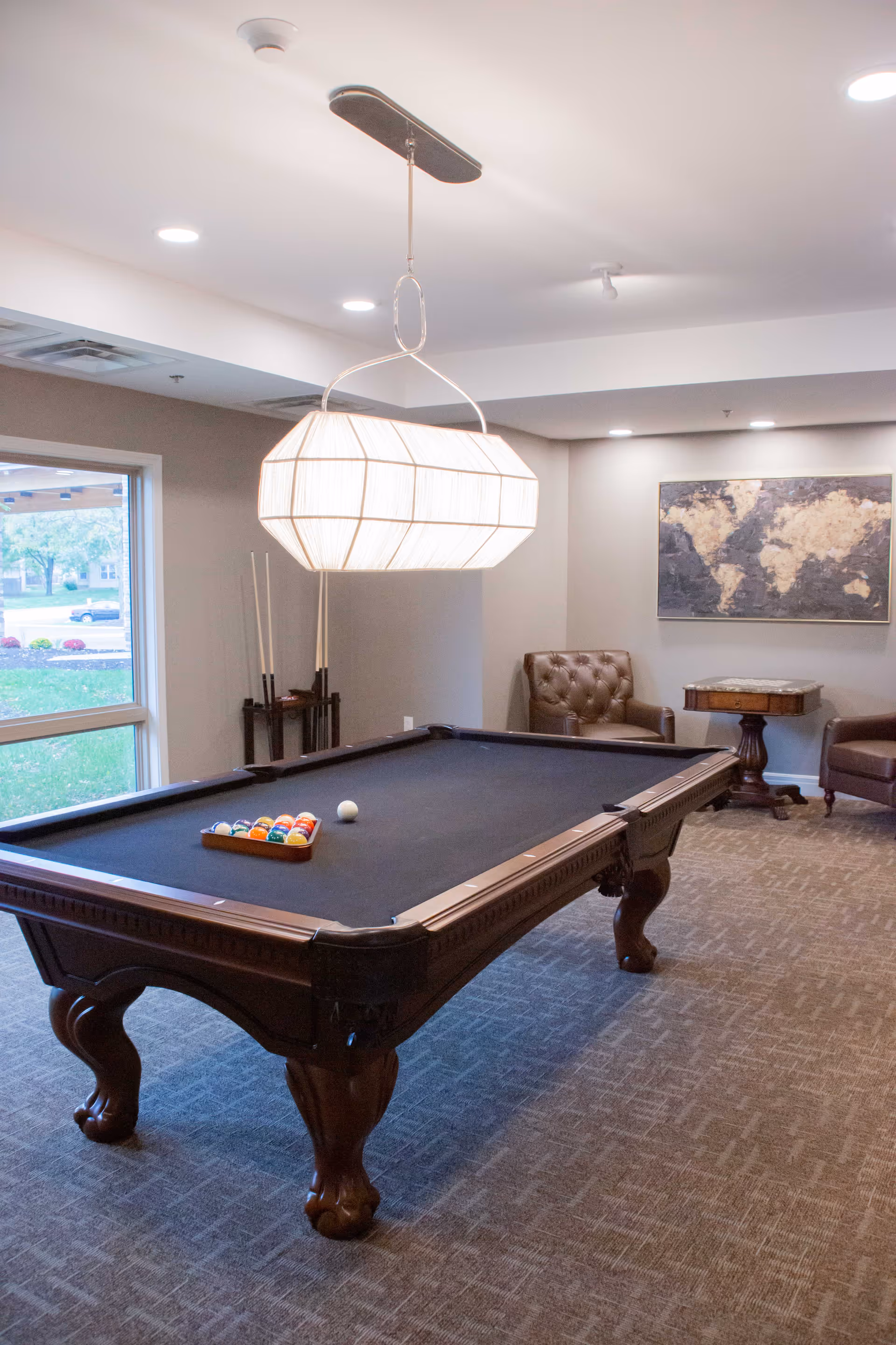 A well-lit game room with a dark wood pool table featuring a black felt surface and a rack of billiard balls. The room has a large window showing greenery outside, a modern hanging light fixture above the pool table, two brown leather armchairs, a small wooden table, and a world map artwork on the wall.