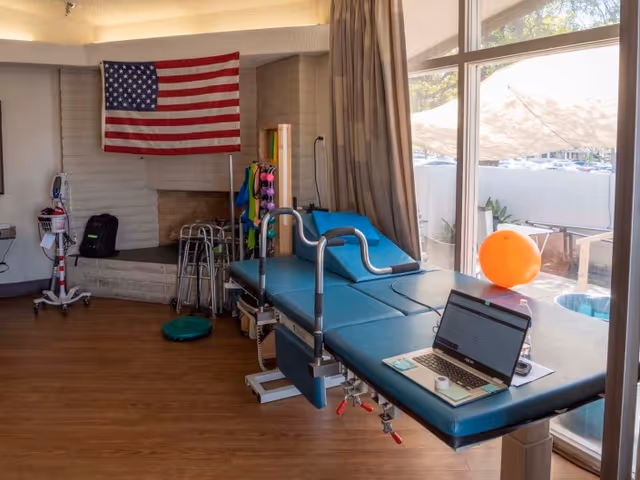 A therapy or rehabilitation room with a blue padded treatment table equipped with metal handrails. A laptop and some small items are placed on the table. The room has wooden flooring, a large window with beige curtains, and an American flag hanging on a white brick wall. Various therapy equipment, including walkers and exercise bands, are visible near the wall.