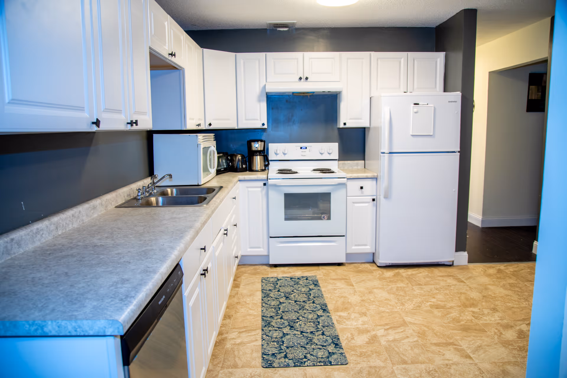 A clean kitchen with white cabinets, a white refrigerator, a white stove with an oven, a microwave, a coffee maker, a toaster, a double sink, and a dishwasher. The walls are painted dark gray, and there is a patterned runner rug on the beige tiled floor.