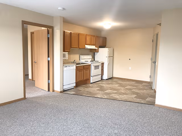 Interior view of a senior living facility apartment showing a small kitchen area with wooden cabinets, a white refrigerator, stove, microwave, and dishwasher. The kitchen floor is tiled, and the adjacent living area is carpeted. There is an open doorway leading to another room.
