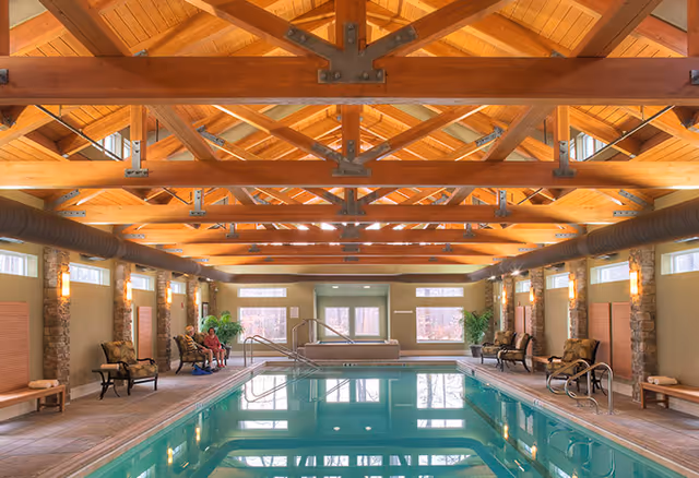 Indoor swimming pool with exposed wooden beams, seating along the sides and a hot tub at the far end.