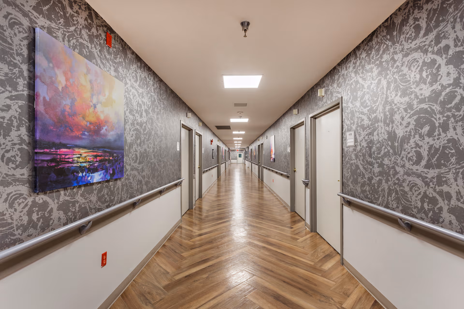 Long indoor hallway with patterned gray wallpaper and wooden herringbone flooring. Several closed doors line both sides of the hallway, each with handrails running along the walls. Bright ceiling lights illuminate the corridor, and colorful abstract paintings are hung on the walls.