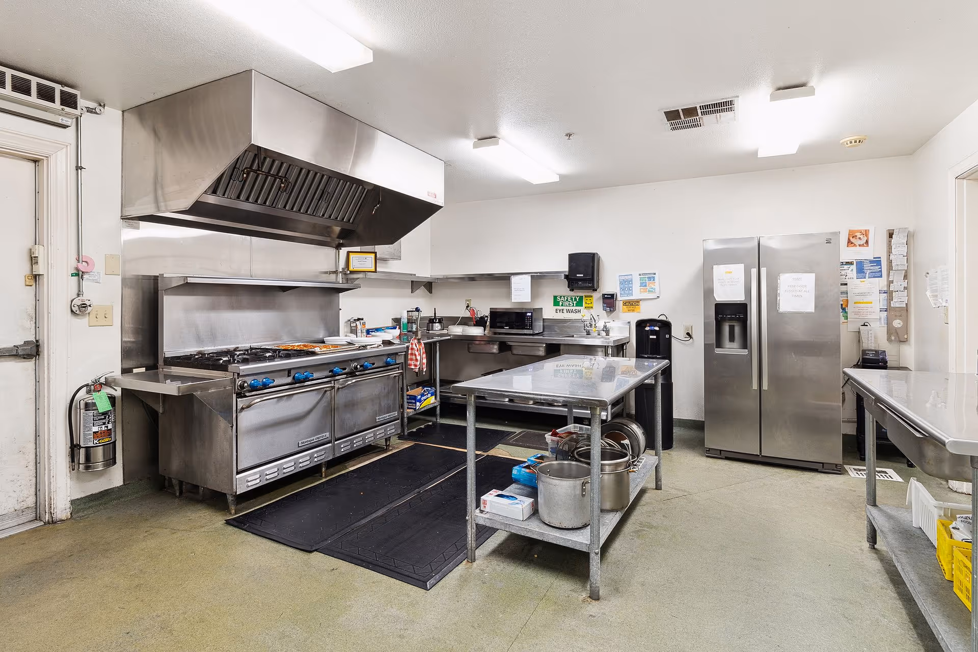 A commercial kitchen with stainless steel appliances including a large stove with multiple burners and an overhead exhaust hood, a double-door refrigerator, stainless steel tables with kitchen utensils and supplies, and various safety and instructional signs on the walls.