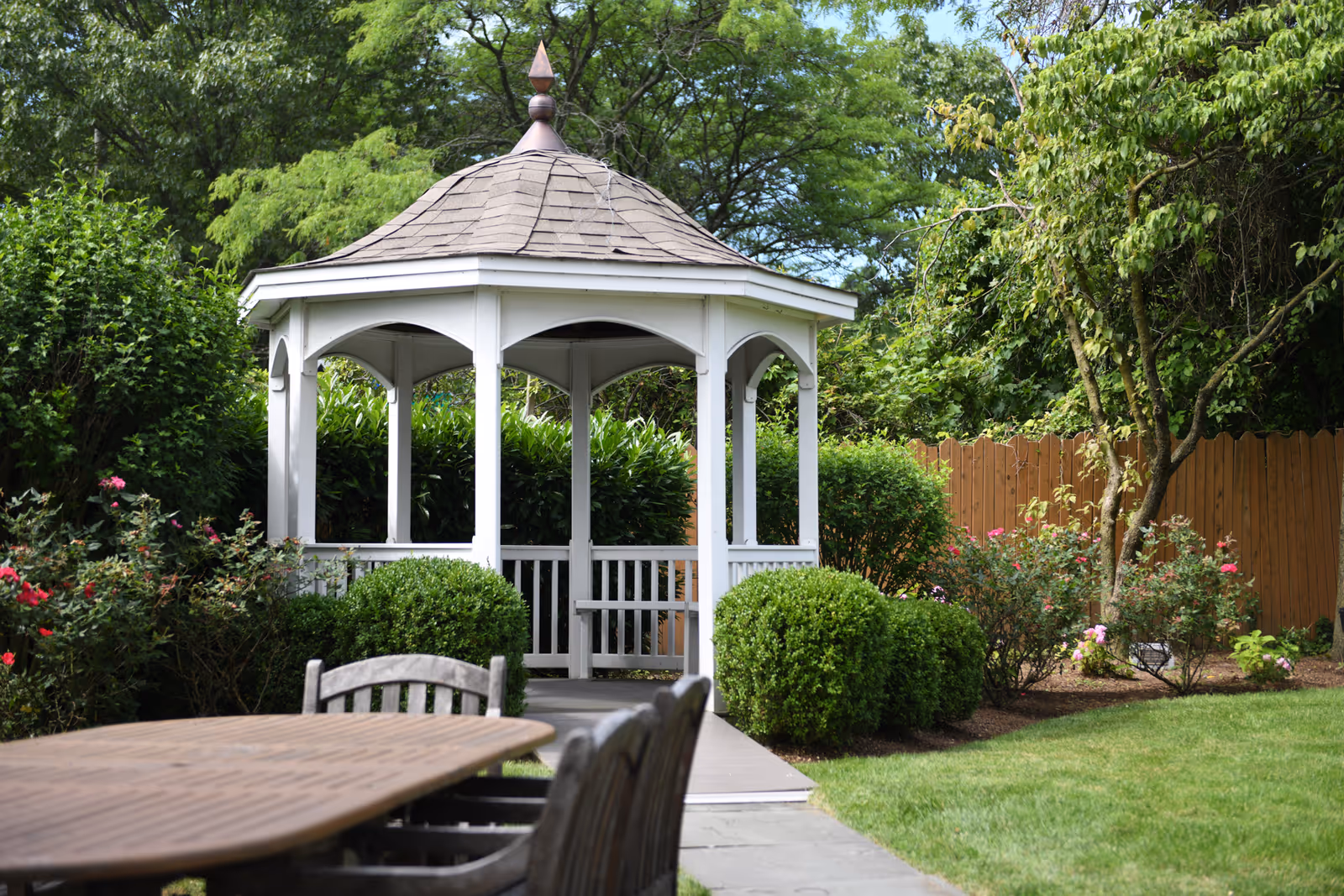 A white wooden gazebo with a shingled roof in a garden surrounded by green bushes, trees, and flowering plants. In the foreground, there is a wooden table with chairs on a stone patio.