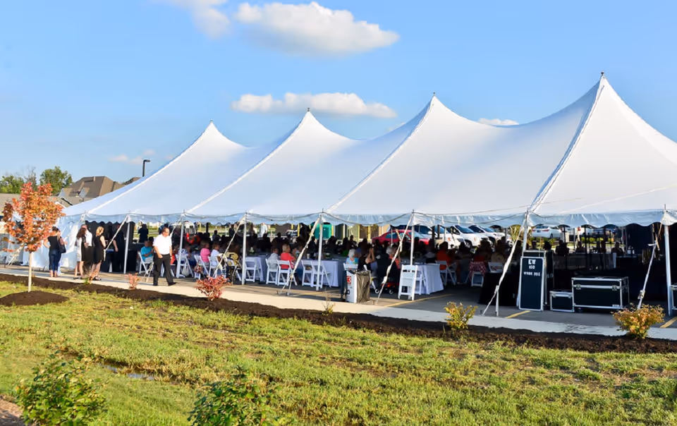 Large white event tent set up outdoors on a sunny day with many people seated inside on white chairs, some standing outside. The tent is on a paved area next to a grassy lawn with small trees and shrubs.