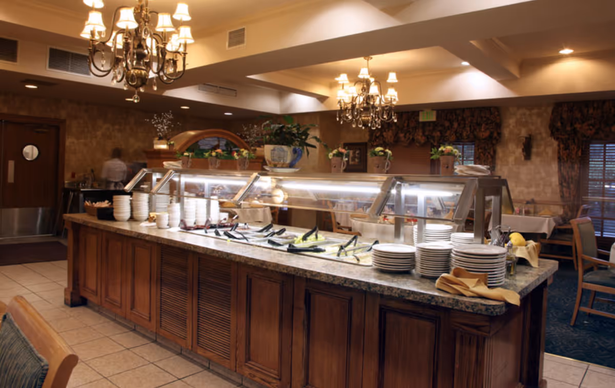 A warmly lit dining room with a long buffet-style serving counter holding stacked plates and a salad bar.