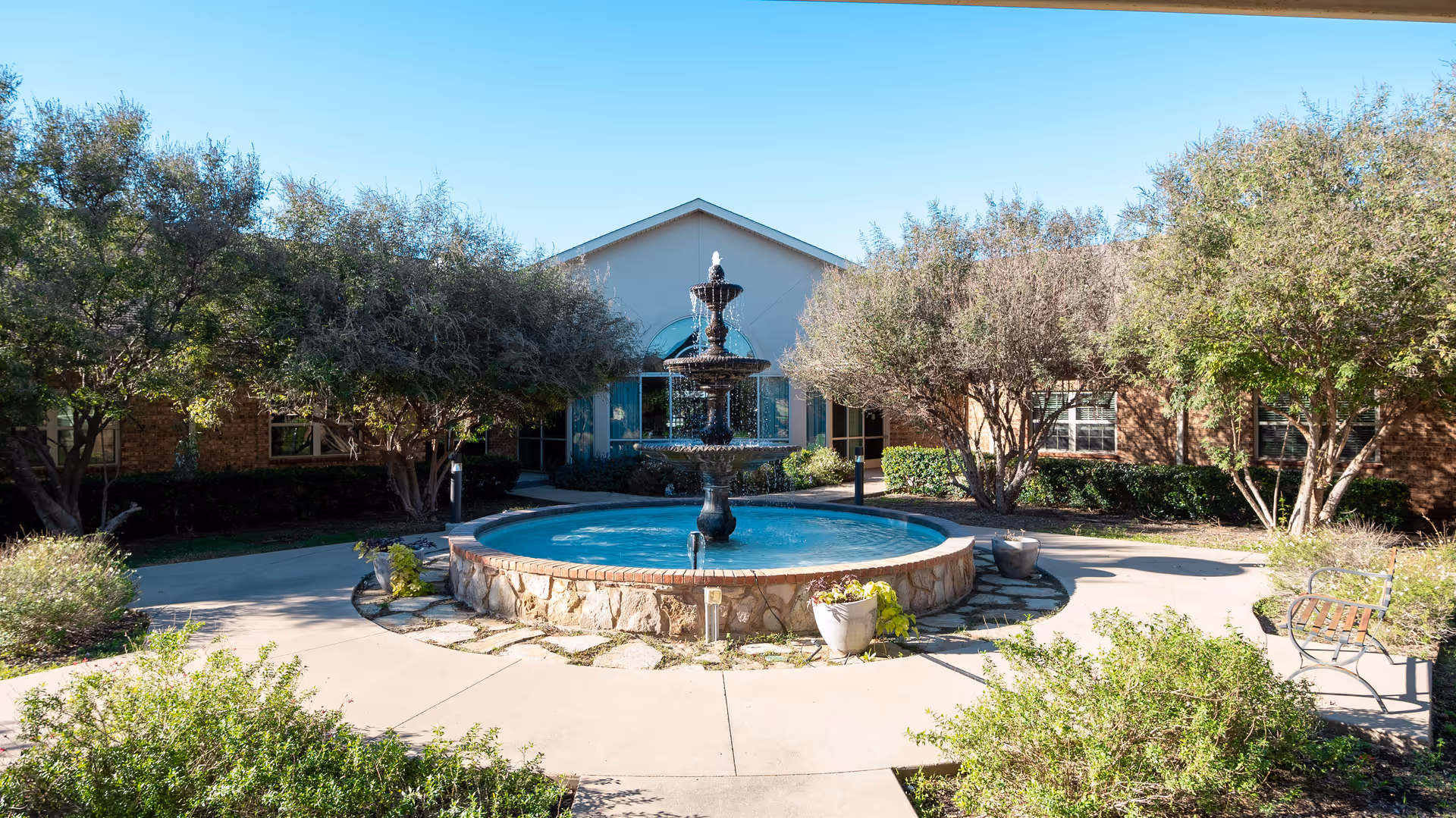 Outdoor courtyard area of Midlothian Healthcare Center featuring a large circular stone fountain with water flowing, surrounded by trees, shrubs, and a paved walkway with benches.