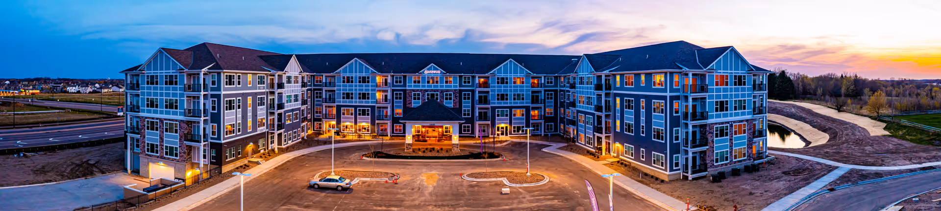 Wide panoramic view of a large, modern senior living facility building at dusk with lights on inside. The building is three stories tall with multiple windows and balconies. There is a circular driveway and parking area in front, with a few cars parked. Surrounding the building are paved roads, landscaped areas, and a small pond to the right.
