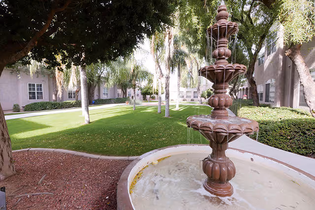 Outdoor courtyard area with a multi-tiered water fountain in the foreground, surrounded by green grass, trees, and a building with multiple windows in the background.